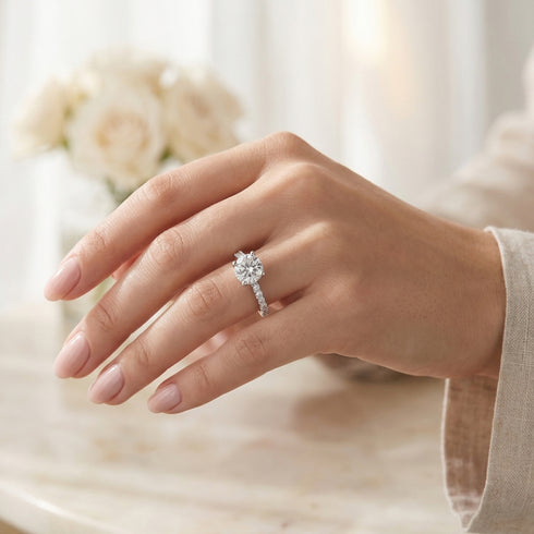A close-up of a hand wearing a diamond engagement ring with a round-cut center stone and a pavé band, set against a softly blurred background with cream roses.